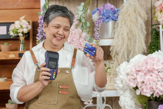 Asian elderly female shop owner standing inside floral boutique smiling while holding credit card and card reader promoting small business accepting digital transactions after retirement