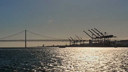 . Silhouettes of cranes and Ponte 25 de Abril bridge along Tagus river against a colorful evening sky. Lisbon, Portugal 