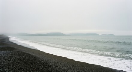 Black pebble beach on a foggy coastline. Gentle ocean waves with white sea foam. Misty mountains on the horizon. Minimalist nature landscape for wellness and tranquility concepts