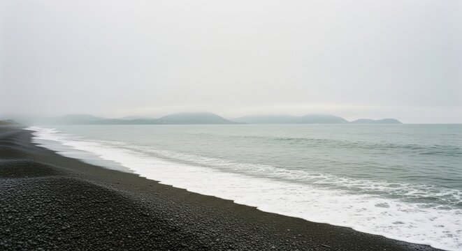 Black pebble beach on a foggy coastline. Gentle ocean waves with white sea foam. Misty mountains on the horizon. Minimalist nature landscape for wellness and tranquility concepts