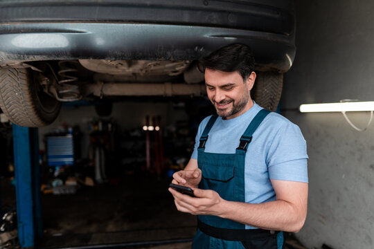 Mechanic smiles while checking smartphone under lifted car in workshop - Powered by Adobe