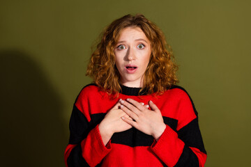 Young redhead woman in red and black striped sweater looks surprised with hands on her chest against khaki background