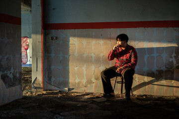 Young Asian man in a plaid shirt sitting and smokes a cigarette in abandoned building alone.