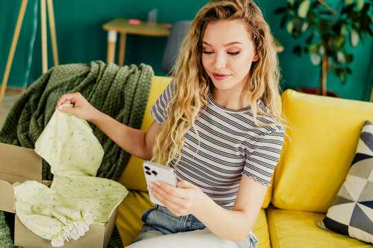 Relaxed young woman at home opening a box of clothes on a yellow couch while checking her phone in a cozy living room - Powered by Adobe