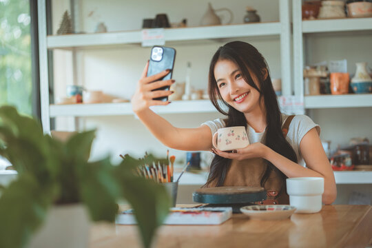 Happy asian female artist taking selfie with handmade pottery design in creative studio – smiling sculptor sharing artwork on social media for online selling, digital portfolio and creative expression - Powered by Adobe