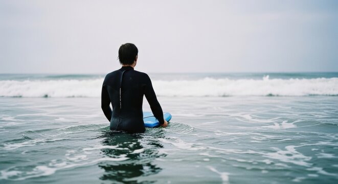 Adult Caucasian male surfer in a black wetsuit holding a blue board. Waiting in the ocean for the perfect wave. Extreme water sports and active lifestyle concept. Personal challenge and solitude
