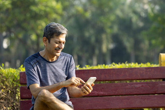 Indian Senior Man Sitting on Park Bench Using Smartphone to Relax After Exercise