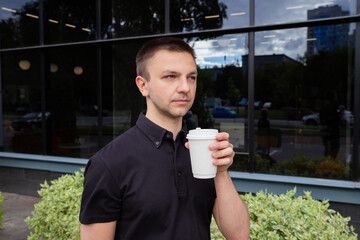 Young Caucasian man enjoys coffee outdoors, sipping coffee from disposable white cup. The scene captures bright day with reflections in the glass surfaces around him, showcasing urban life