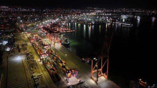 Nighttime overhead view of gantry cranes unloading containers at mersin international port, brightly lit terminal against the city skyline and dark mediterranean sea