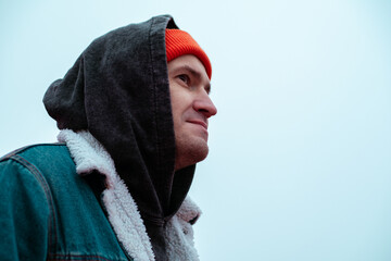 A young man stands outdoors in a casual outfit featuring a hoodie and beanie He gazes upwards seemingly lost in thought against a grey overcast sky