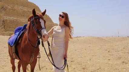 Woman enjoying a vacation in Egypt, standing beside a horse near ancient pyramids in the desert under the bright sun.