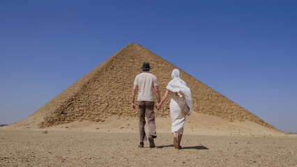 Couple holding hands and walking toward the pyramid in Egypt, enjoying a sunny vacation and cultural travel experience.
