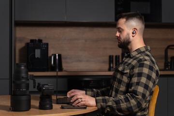Male photographer or videographer editing content on a laptop, sitting at a table with camera equipment in a modern home.