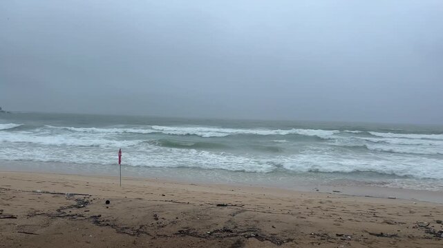 Stormy sea with large, powerful waves crashing on a sandy beach. Red warning flag indicating dangerous swimming conditions during bad weather