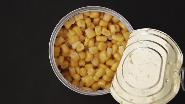 Black background top view of opened metallic tin exposing yellow corn kernels arranged in clear composition for preserved ingredient display