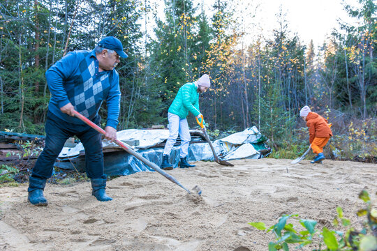 Three people working together outdoors in a forested area. They are shoveling sand. The scene shows a sunny day with trees in the background.