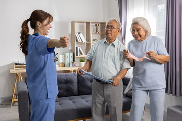 Asian Elderly couple doing resistance band exercises with female caregiver in cozy living room,...