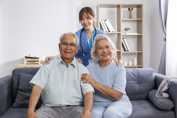 Fototapeta premium Asian Elderly couple smiling on sofa with female caregiver behind them offering support and warmth in bright living room