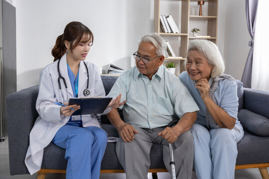 Asian young female doctor consulting elderly couple about health plan and medication on sofa with clipboard and stethoscope, caring medical discussion, supportive home visit - Powered by Adobe