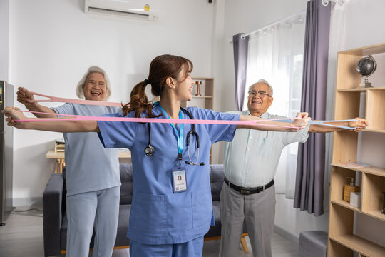 Asian female healthcare worker leading senior woman and senior man in resistance band exercise at home, physical therapy, rehabilitation, home care, smiling caregiver