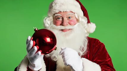 Happy Santa Claus holds red ornament on green screen backdrop, spreading holiday cheer and festive spirit with a joyful, classic Christmas scene