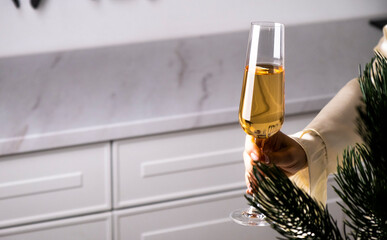 Glass of sparkling wine in woman hand at kitchen background near Christmas tree branch. Cozy, elegant and festive atmosphere. Winter holidays celebration. Merry Christmas and Happy New Year.