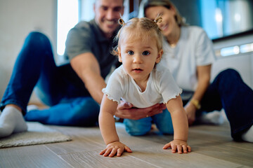 Baby girl crawls across the living room floor while mother and father gently support and encourage her first developmental milestones at home