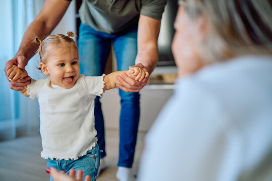 Toddler taking first steps, holding parents' hands, with a joyful expression, while learning to walk at home
