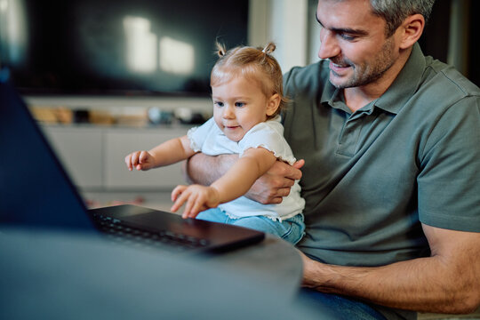 Happy father holding baby daughter, both smiling and interacting with a laptop for family bonding, technology, and parenthood concepts - Powered by Adobe