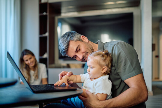 Father and infant daughter are interacting with a laptop at home, with mother in the background. Family bonding and digital learning