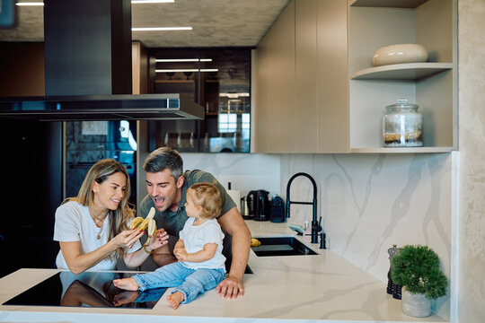 Happy family spending time in a modern kitchen, mother feeding a banana to the father while their child is sitting on the counter - Powered by Adobe