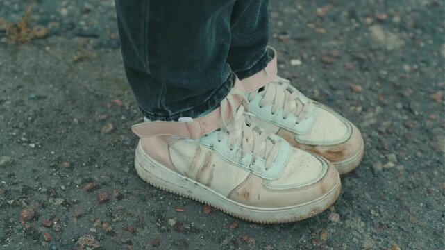 A person stands on a gravel path wearing dirty white sneakers The shoes are scuffed and stained indicating outdoor activities The surroundings are slightly muddy and dull