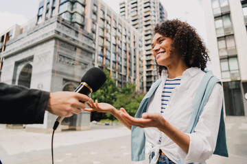 Smiling woman in striped shirt being interviewed on an urban street with microphone reporter and modern buildings in the background © deagreez
