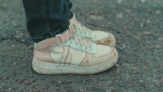 A person stands on a gravel path wearing dirty white sneakers The shoes are scuffed and stained indicating outdoor activities The surroundings are slightly muddy and dull