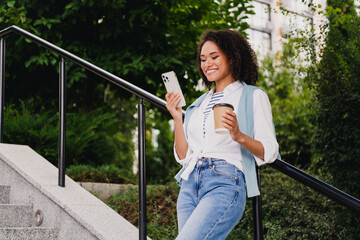 Young woman walks down outdoor stairs smiling while holding coffee and phone in a sunny urban park setting and enjoying a casual city walk