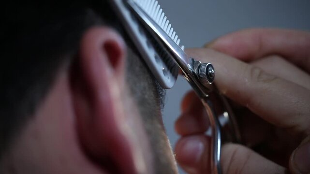 A professional barber uses sharp scissors and a comb for precise hair trimming around the ear. Close-up detail of the salon cutting process.
