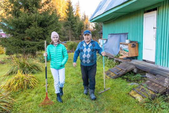 A young Caucasian woman and a senior Caucasian man walk in a garden. They hold gardening tools and wear casual outdoor clothing. A green house is in the background.