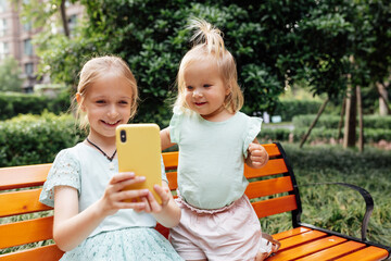 Two young girls sitting on a bench taking a selfie with a smartphone in a park