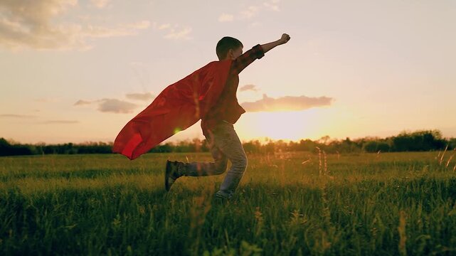 Powerful superhero boy in red cloak running on sunset field with victory gesture side view slowmo. Brave male teen in hero costume fantasy flying imagination winner protection playing game at meadow
