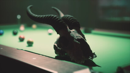 Fototapeta premium In a dimly lit room, a goat skull rests on the edge of a vibrant green pool table surrounded by colorful billiard balls. The eerie ambiance adds a touch of mystery to the scene.