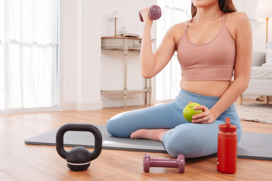 Fit woman resting after home workout, sitting on yoga mat with kettlebell, water bottle, and green apple healthy lifestyle, self-care, and recovery. - Powered by Adobe