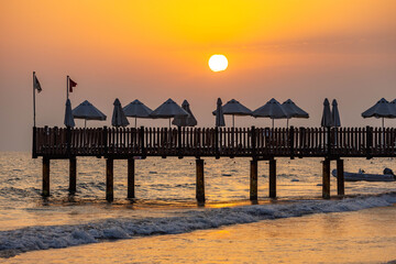 Wooden sundeck over sea with sunbed and umbrella silhouettes against orange sunset sky, large sun disk reflecting on sparkling water with dinghy below. Mediterranean resort luxury.