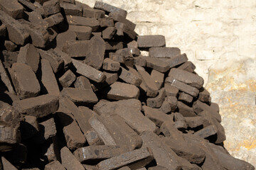 A large pile of peat briquettes is shown stacked neatly, showcasing their dark brown color, against an old, textured wall