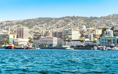 Colorful coastal city skyline with boats and hills under clear sky, vibrant waterfront urban landscape and harbor view.