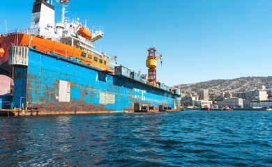 Large dry dock with crane and rusting ship hull in shipyard, industrial construction and maritime repair services, vibrant blue sky and coastal city view.