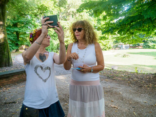 Obraz premium Two women friends capturing memories together outdoors in a park with a smartphone taking a selfie photo with a casual vibe and sunny summer day atmosphere enjoying their time