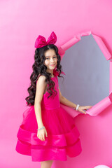 A young Hispanic girl with long, curly black hair wears a bright pink dress and a large pink bow. She stands beside a pink-framed mirror against a pink background.