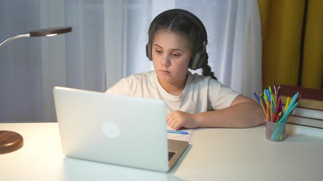 Young girl wearing headphones and sitting at desk with laptop. Young girl is learning lifestyle how to use laptop in distance. Child is studying at a table during evening lessons. A young girl is.