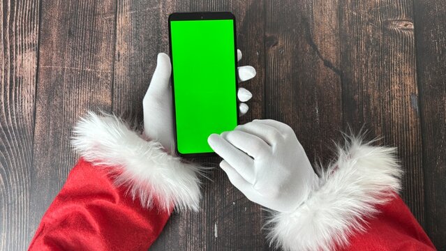  Festive Santa hand in a red and white costume holds a smartphone with a vibrant green screen. Perfect for displaying Christmas-themed digital content