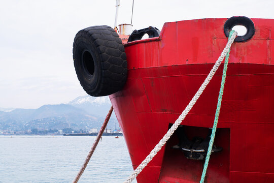 The bow of a red ship with a raised anchor, mooring of the vessel.
An old car tire is used as a buffer between the ship and the pier. The ship is secured with thick mooring ropes.